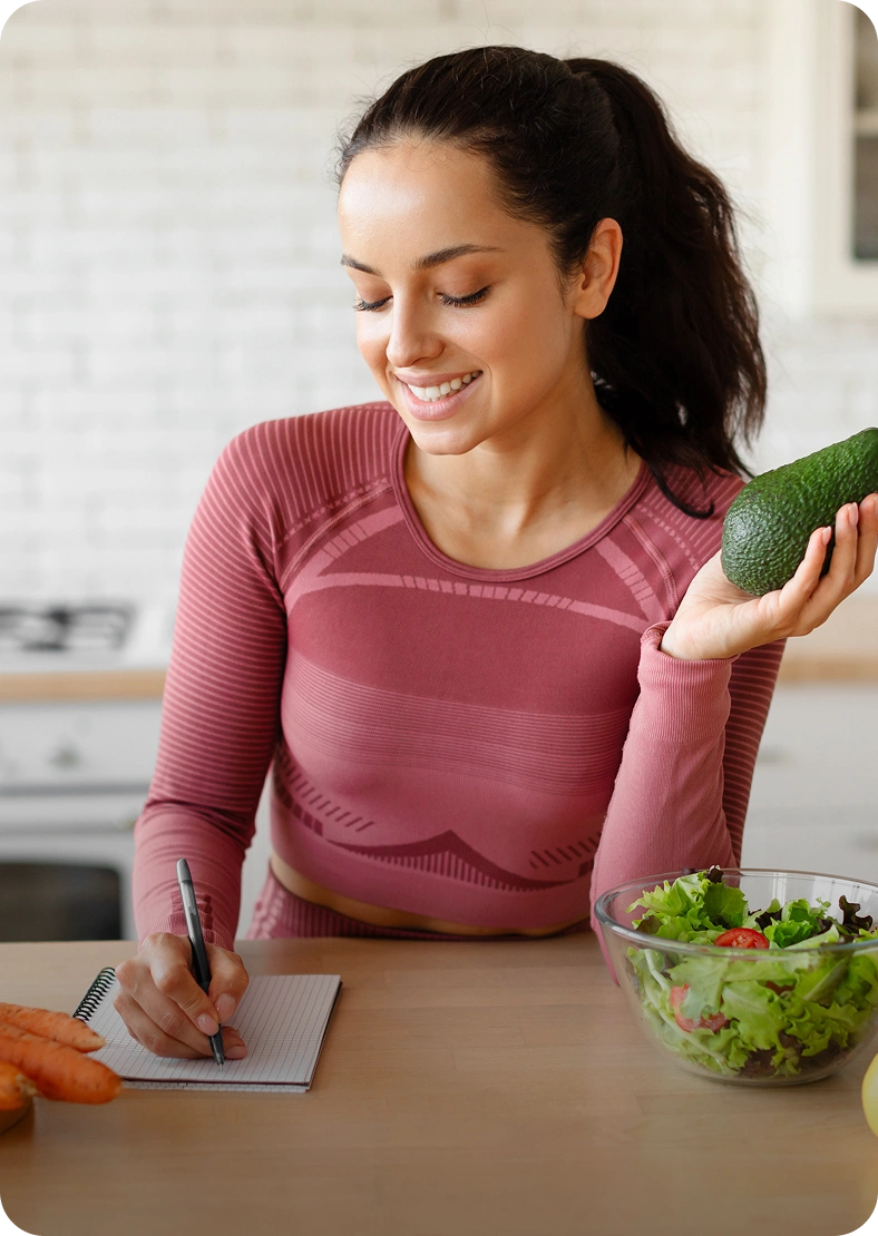 Woman writing, holding avocado, with salad bowl.
